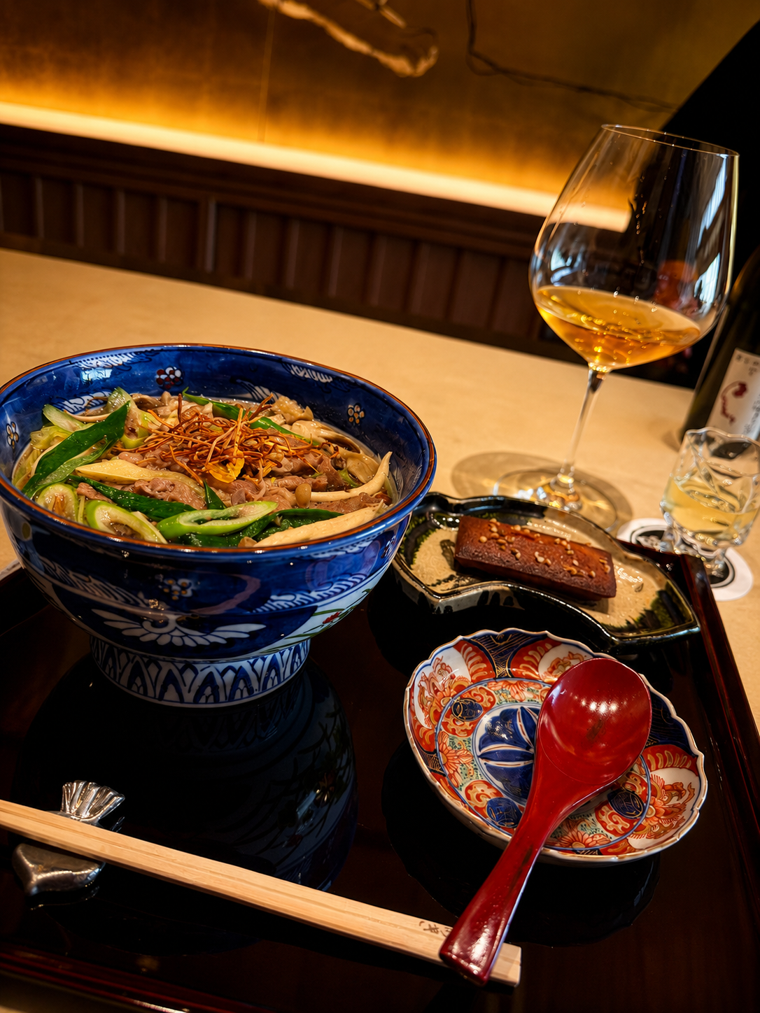 A refined ramen lunch at Saryo Tesshin: a blue-and-white bowl of noodles with greens, a glass of white wine, small Imari dishes on a dark tray