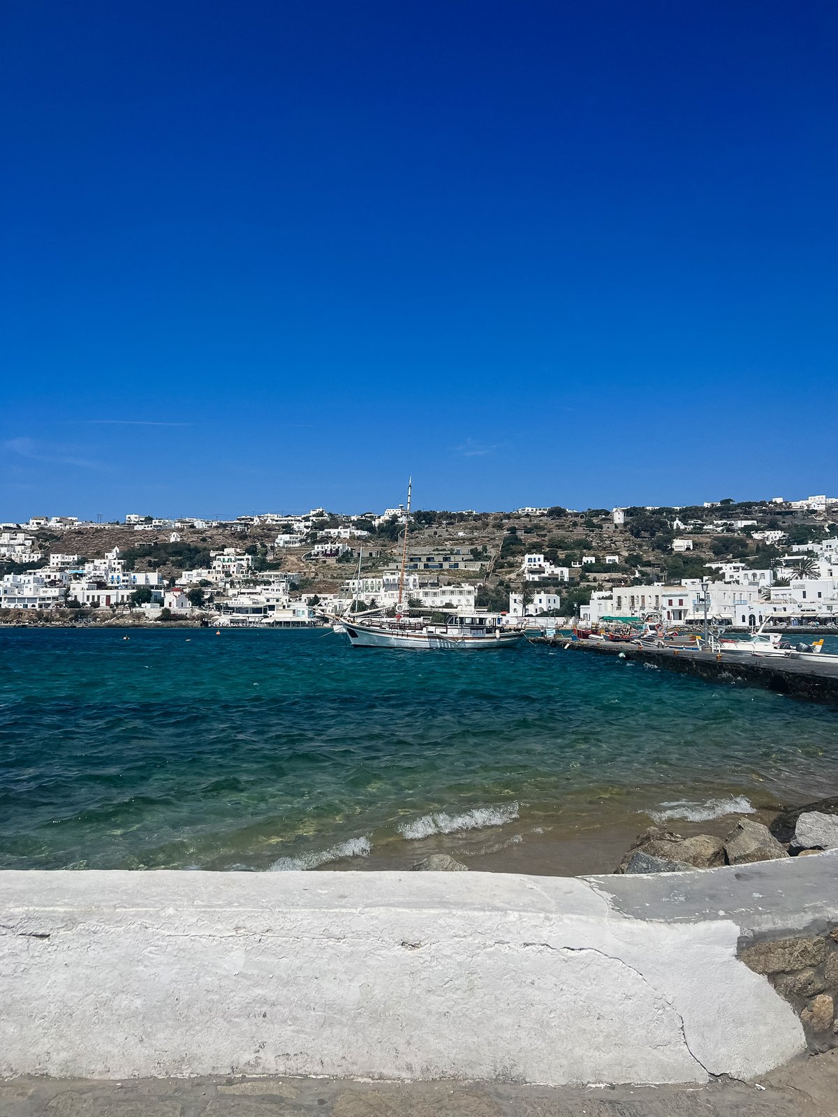 Bright midday view across Mykonos harbor to the whitewashed town climbing the hill, a wooden caique moored in calm blue water