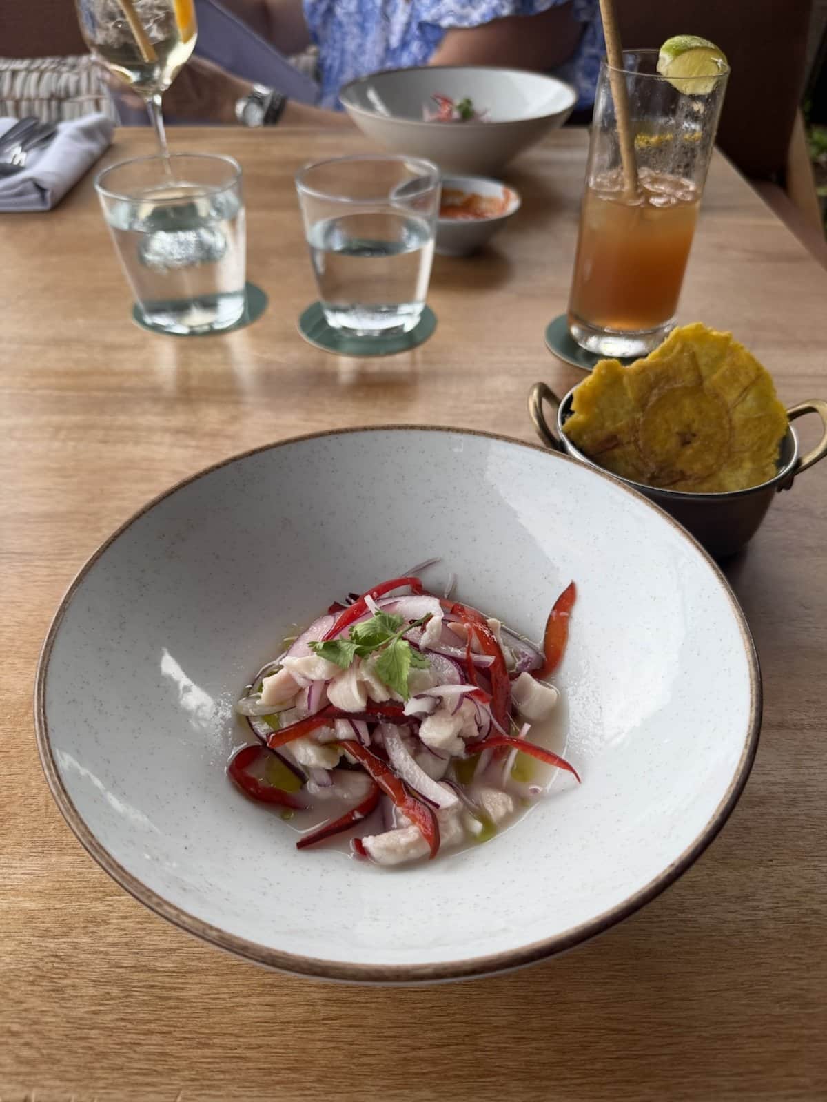 A bowl of Costa Rican ceviche with red onion and peppers, a basket of patacones and an iced drink on a wooden table
