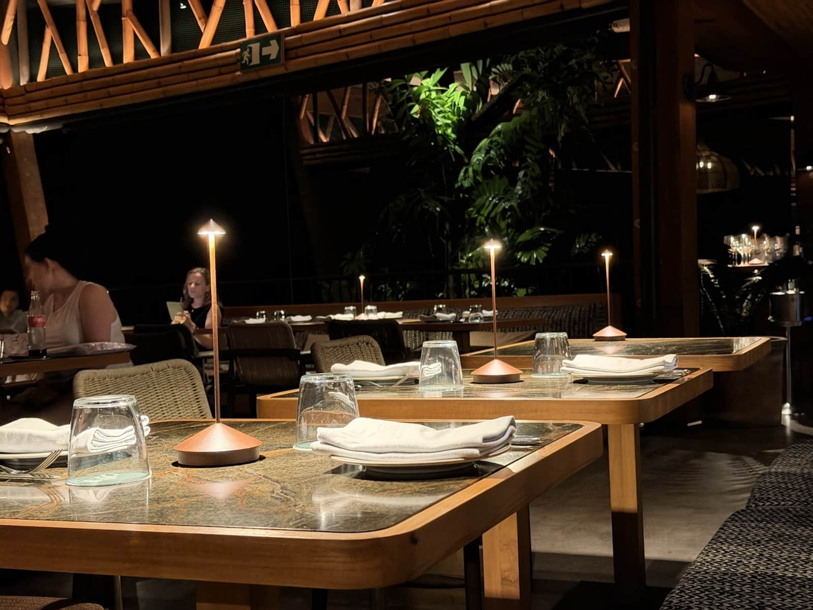 The Gaio dining room at night, set tables with small candle lamps, warm wood ceiling and dark forest beyond the open sides