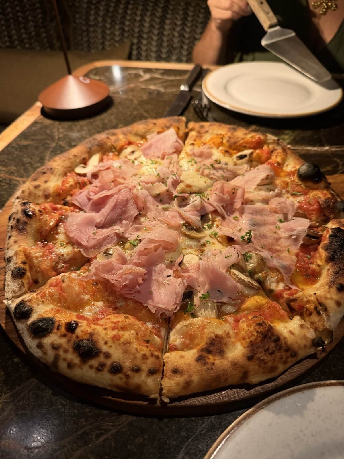 A wood-fired pizza topped with prosciutto cotto and mushrooms on a dark marble table